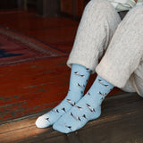 Person sitting indoors on a wooden floor wearing cream knit trousers and baby blue puffin print crew socks.
