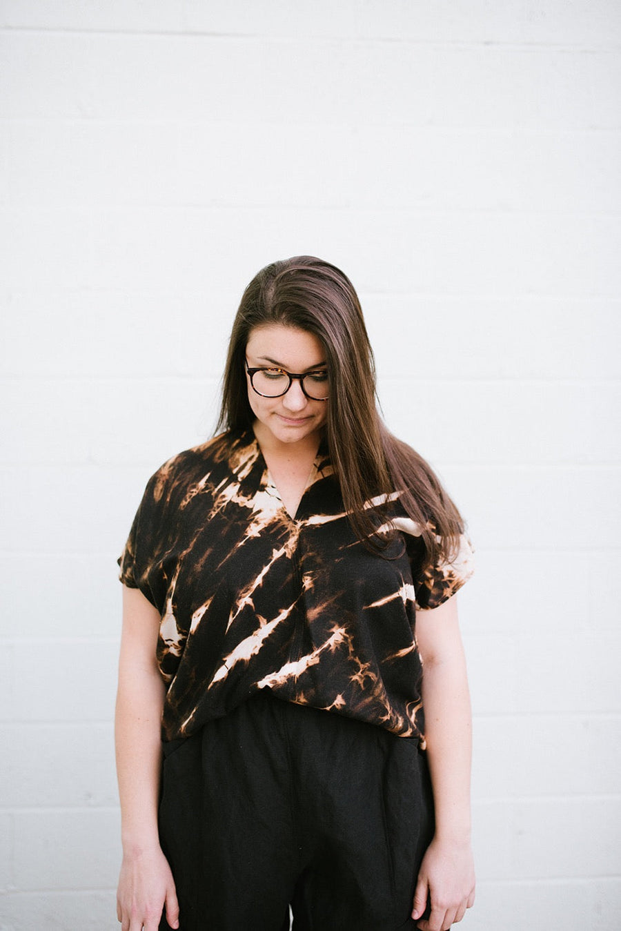 Front view portrait of model Rosemary wearing the black and tan tie-dyed Megan-Ilene Zero Waste Silk Market Top against a white brick wall.