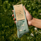 Person holding a packaged pair of Conscious Step Socks That Save Sheep with Farm Sanctuary tag against a natural green field backdrop.