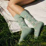 Close-up view of someone lying on a white blanket on grass wearing green Conscious Step Valais Blacknose sheep print socks.