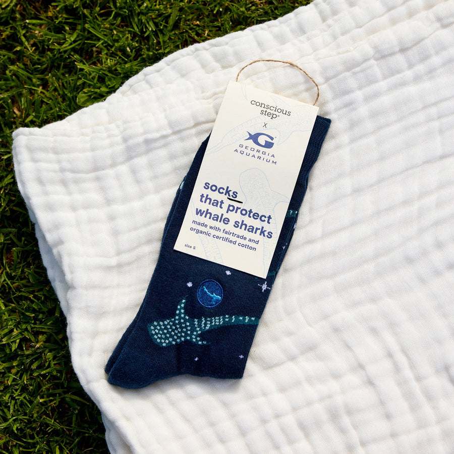 A single packaged pair of navy Conscious Step whale shark socks with a Georgia Aquarium tag, lying on a white crinkled cotton blanket over green grass.