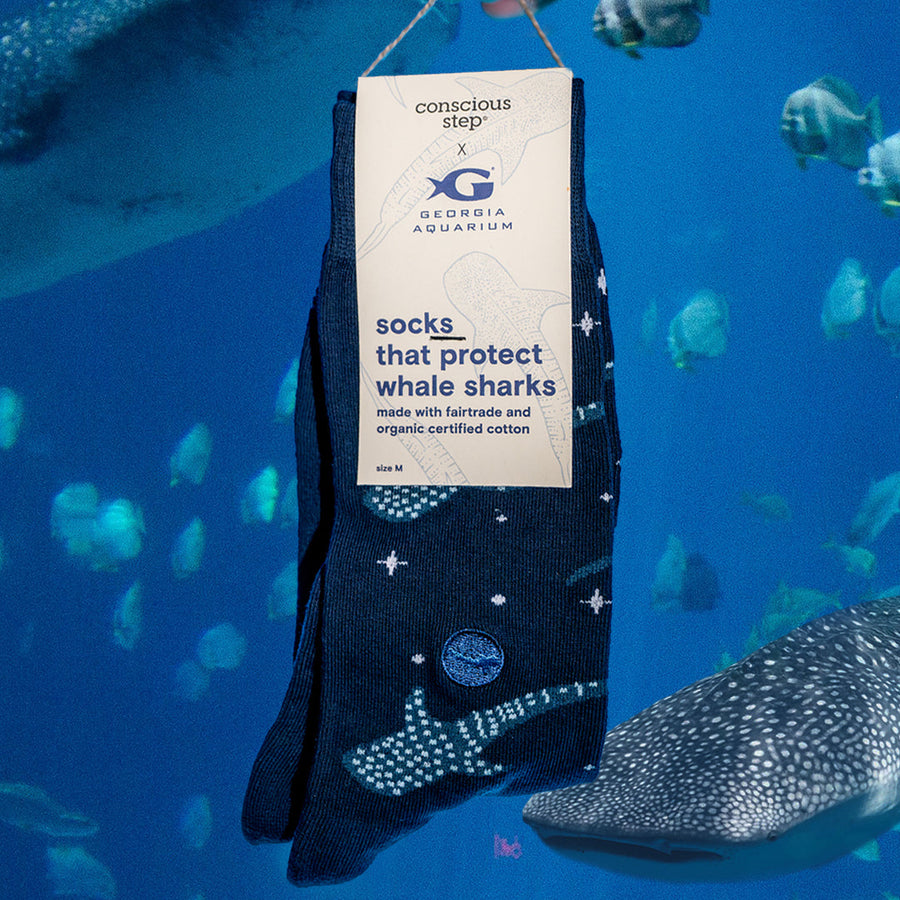 A packaged pair of Conscious Step navy whale shark socks featuring a "Georgia Aquarium" collaboration card sleeve, superimposed over a deep blue underwater ocean scene with swimming whale sharks and fish.