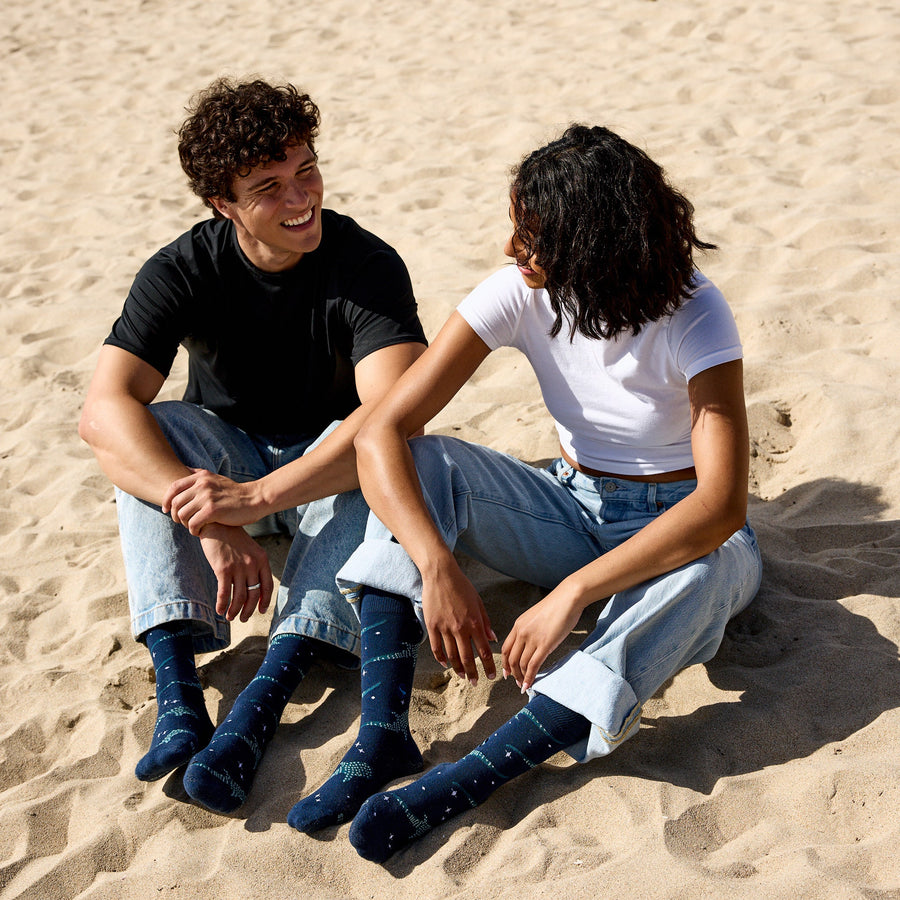 A smiling man in a black t-shirt and a woman in a white t-shirt sitting together on a sandy beach, wearing cuffed blue jeans and matching navy Conscious Step whale shark socks.