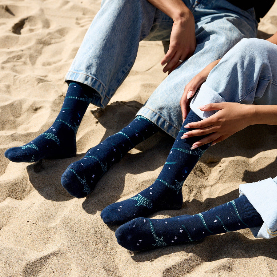 A close-up of the feet and lower legs of two people sitting on beach sand in rolled-up light-wash denim jeans, showcasing their matching navy Conscious Step whale shark crew socks.