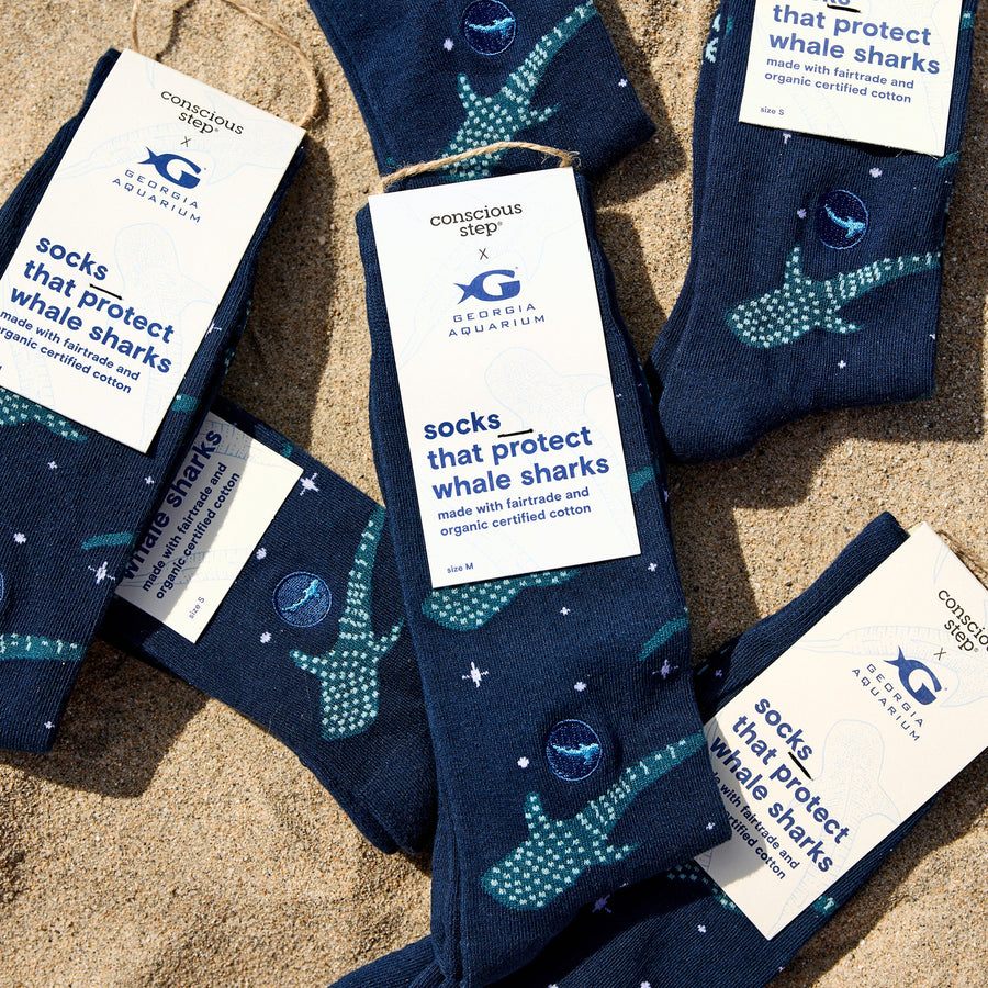 Multiple pairs of folded Conscious Step navy whale shark socks with "Georgia Aquarium" packaging tags scattered on the sand.