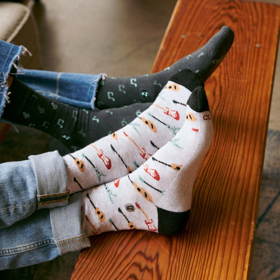 Close-up view of two people relaxing with feet up on a wooden bench, one wearing grey guitar socks and the other wearing dark music note socks.