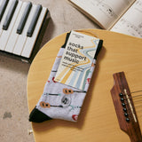 Packaged pair of Conscious Step Socks That Support Music resting on an acoustic guitar next to a piano keyboard and sheet music.