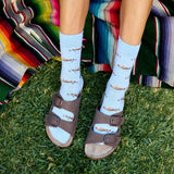 Top-down view of a person's legs and feet on green grass wearing light blue Conscious Step socks with a floating sea otter pattern paired with dark brown two-strap slide sandals, positioned next to a colorful striped blanket.