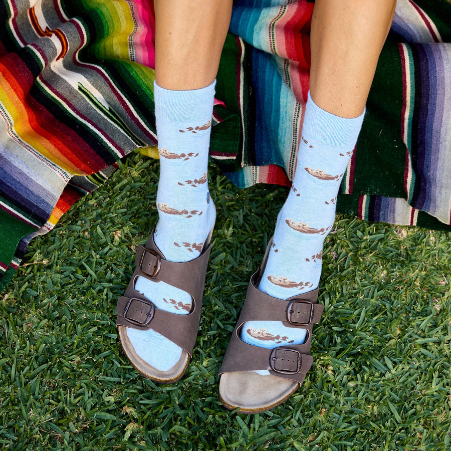 Top-down view of a person's legs and feet on green grass wearing light blue Conscious Step socks with a floating sea otter pattern paired with dark brown two-strap slide sandals, positioned next to a colorful striped blanket.
