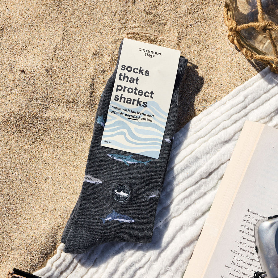 A flat lay on beach sand featuring a packaged pair of dark gray Conscious Step shark socks, an open book, a retro camera, sunglasses, a white beach towel, and a decorative net-wrapped glass bottle.