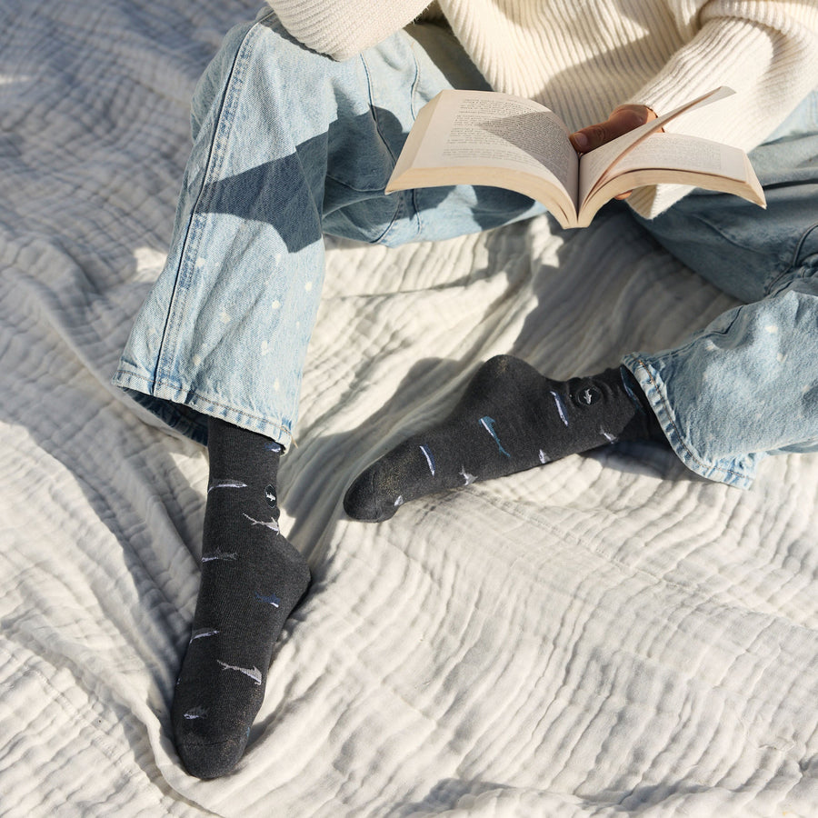 A person sitting on a white blanket wearing light-wash cuffed denim jeans and dark gray Conscious Step shark socks while holding an open book.