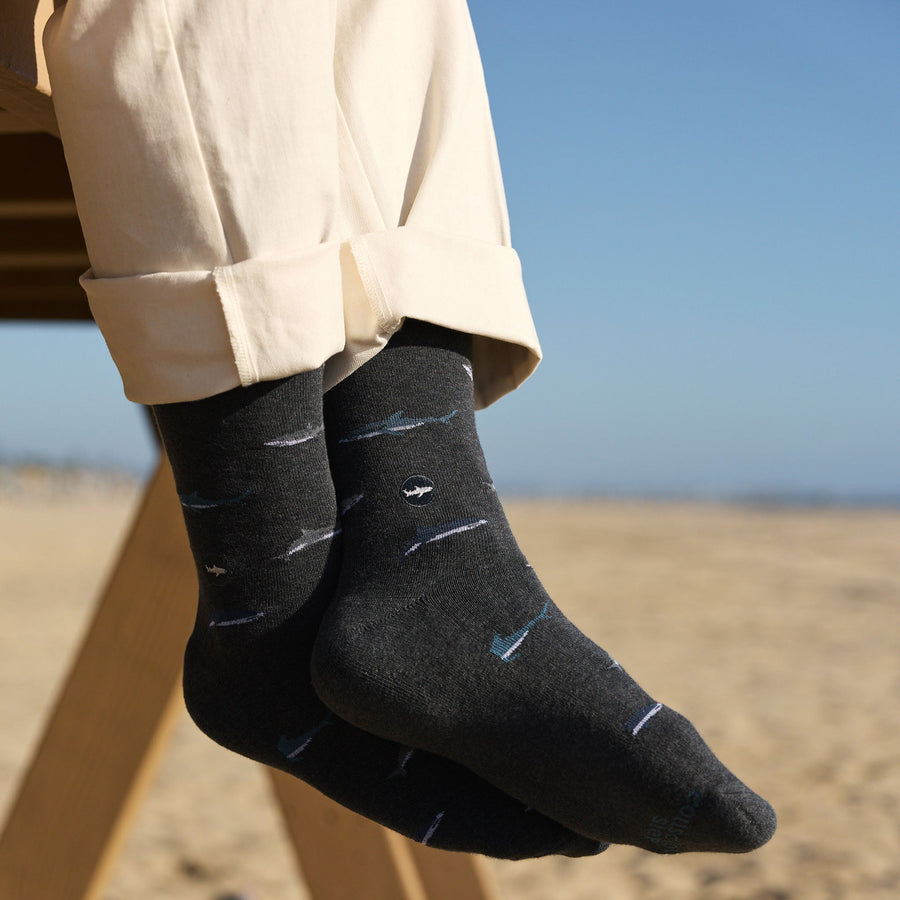 A close-up of a person's ankles and feet dangling from a wooden structure, showing cuffed cream trousers and dark gray crew socks with a repeating shark pattern.