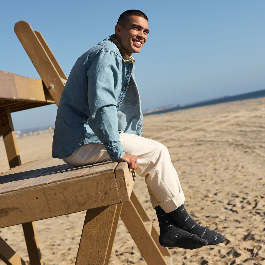 A man in a blue denim shirt and cream trousers sitting on a wooden beach lifeguard tower, smiling and showcasing his dark gray Conscious Step shark socks.