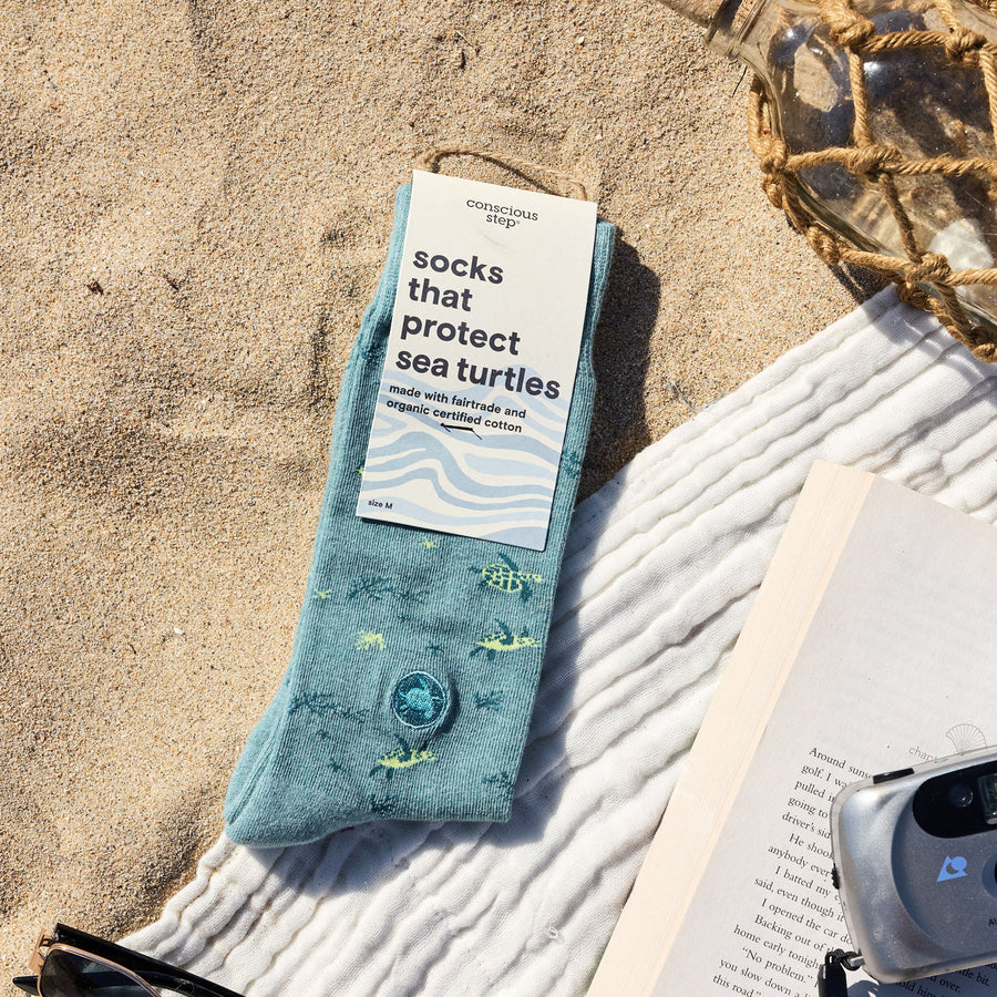 A flat lay on beach sand featuring packaged Conscious Step teal sea turtle socks, an open book, a retro camera, sunglasses, a white beach towel, and a decorative net-wrapped glass bottle.