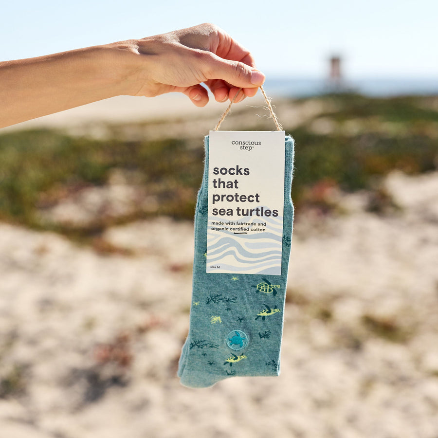 A hand holding a single packaged pair of teal Conscious Step sea turtle socks against a bright, sunny beach background with sandy dunes and a blurred lifeguard tower in the distance.