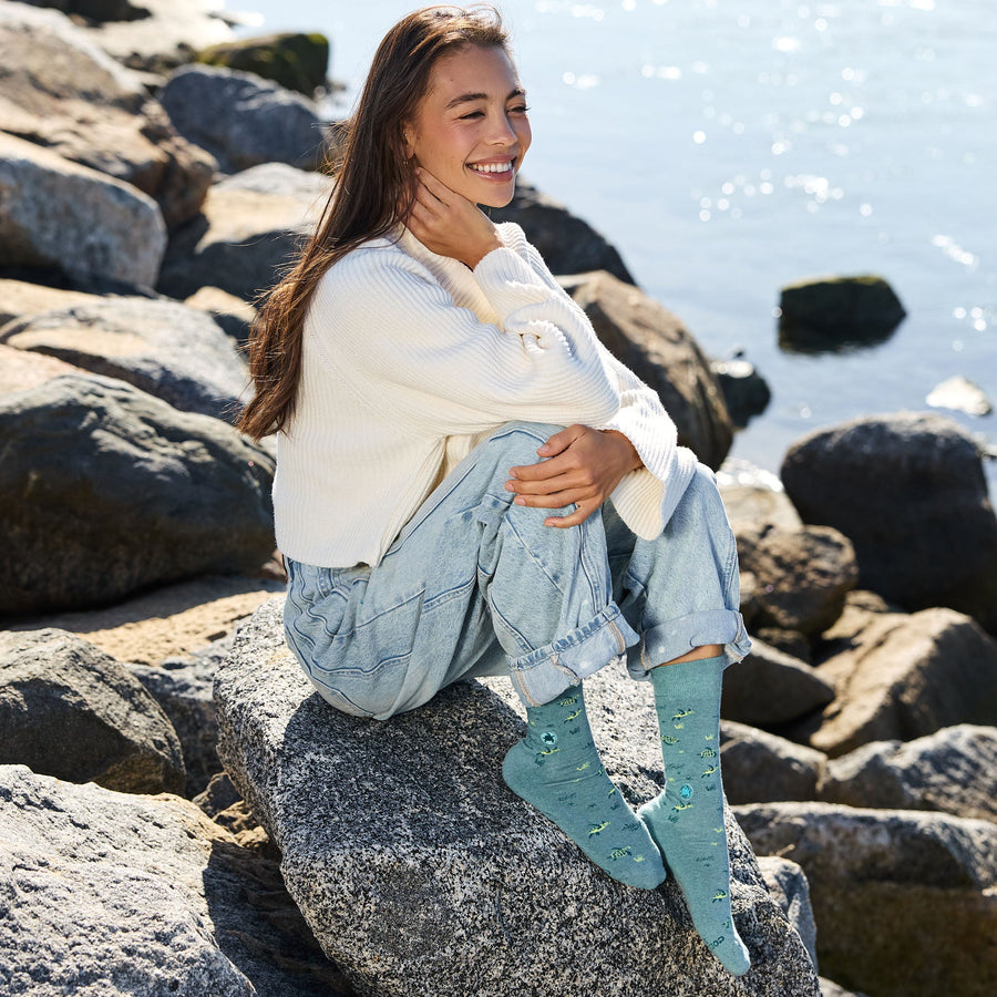 A smiling woman sitting on a large seaside boulder, wearing a cozy white knit sweater, light-wash denim jeans, and light teal Conscious Step sea turtle socks, with the sparkling ocean in the background.