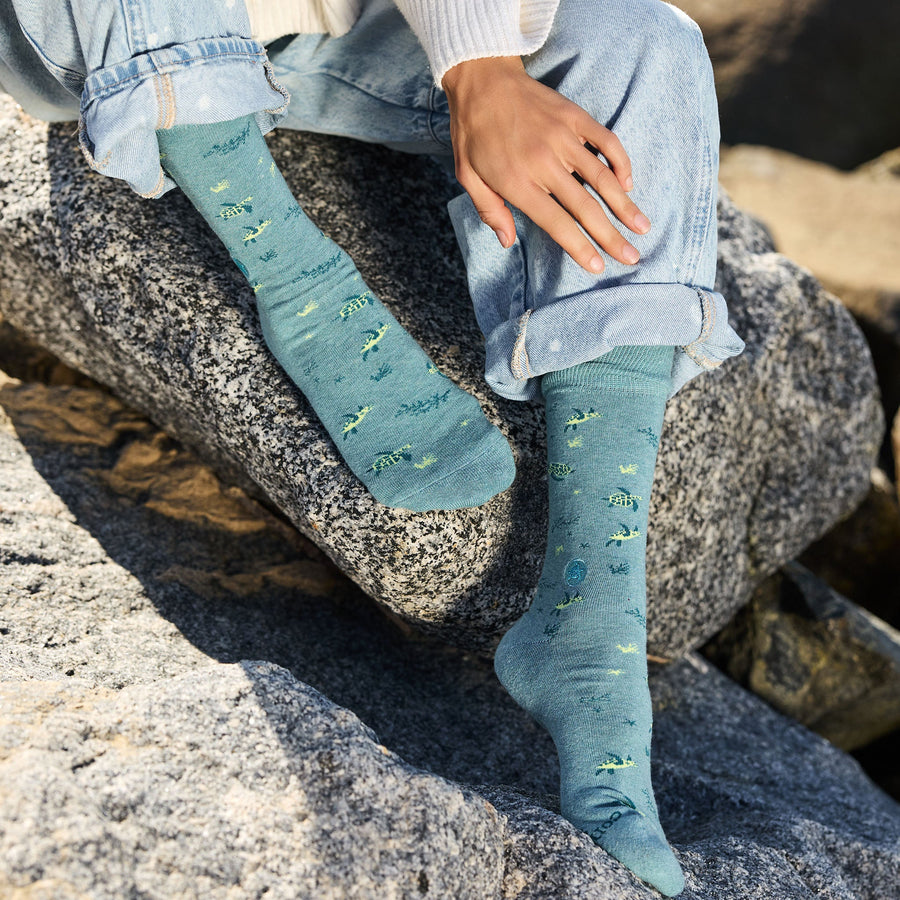 Close-up of a person's feet resting on a dark coastal rock, wearing light-wash jeans and light teal crew socks patterned with small sea turtles and underwater plant designs.