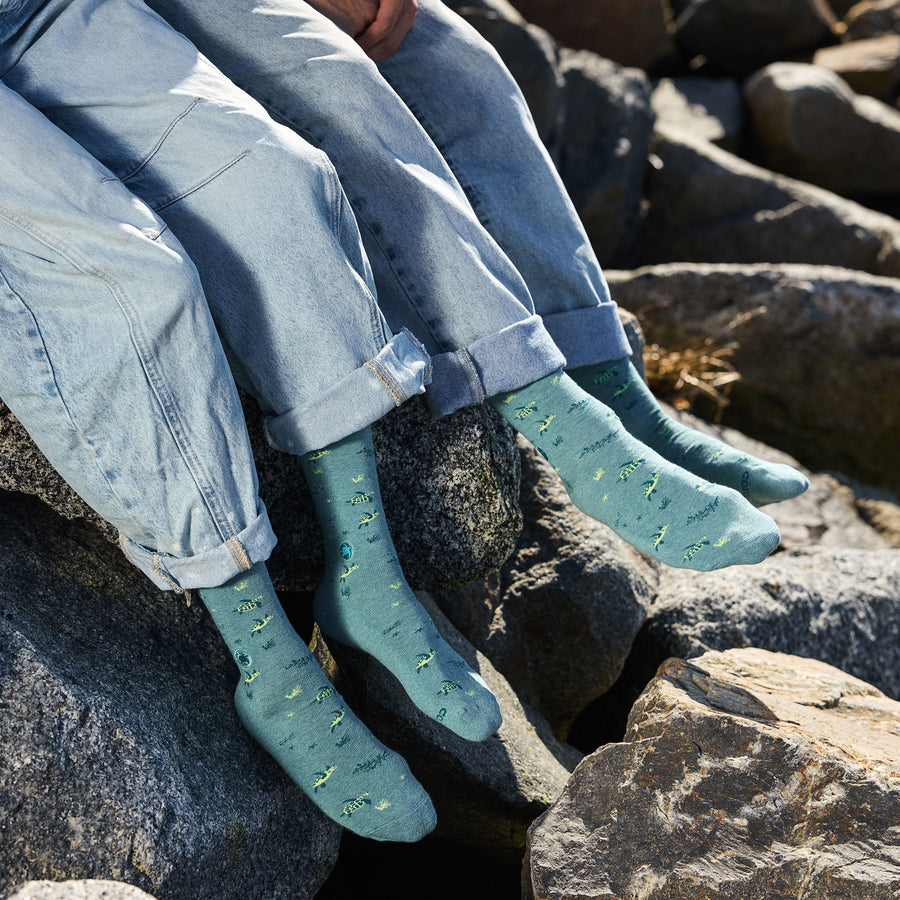A close-up shot of two people sitting side-by-side on dark rocks, showing their lower legs in light-wash denim jeans and wearing matching teal Conscious Step sea turtle crew socks.
