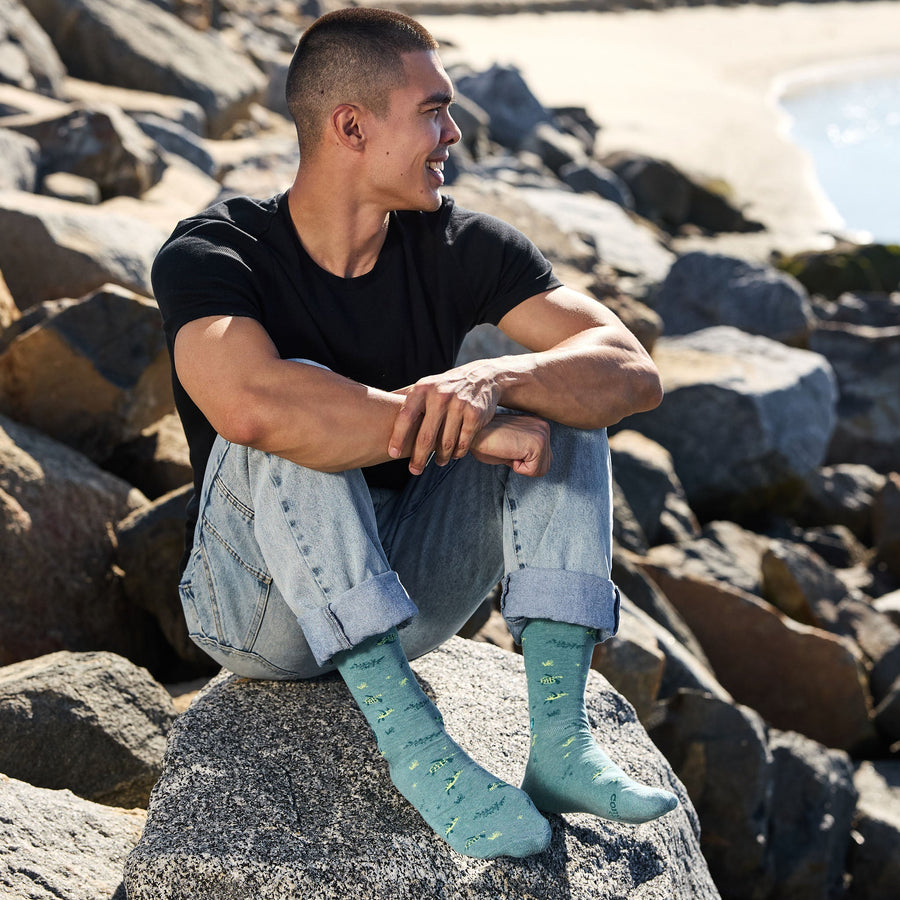 A smiling man in a black t-shirt and rolled-up light-wash jeans sitting on a large coastal rock, showcasing his teal Conscious Step sea turtle socks against a rocky beach background.