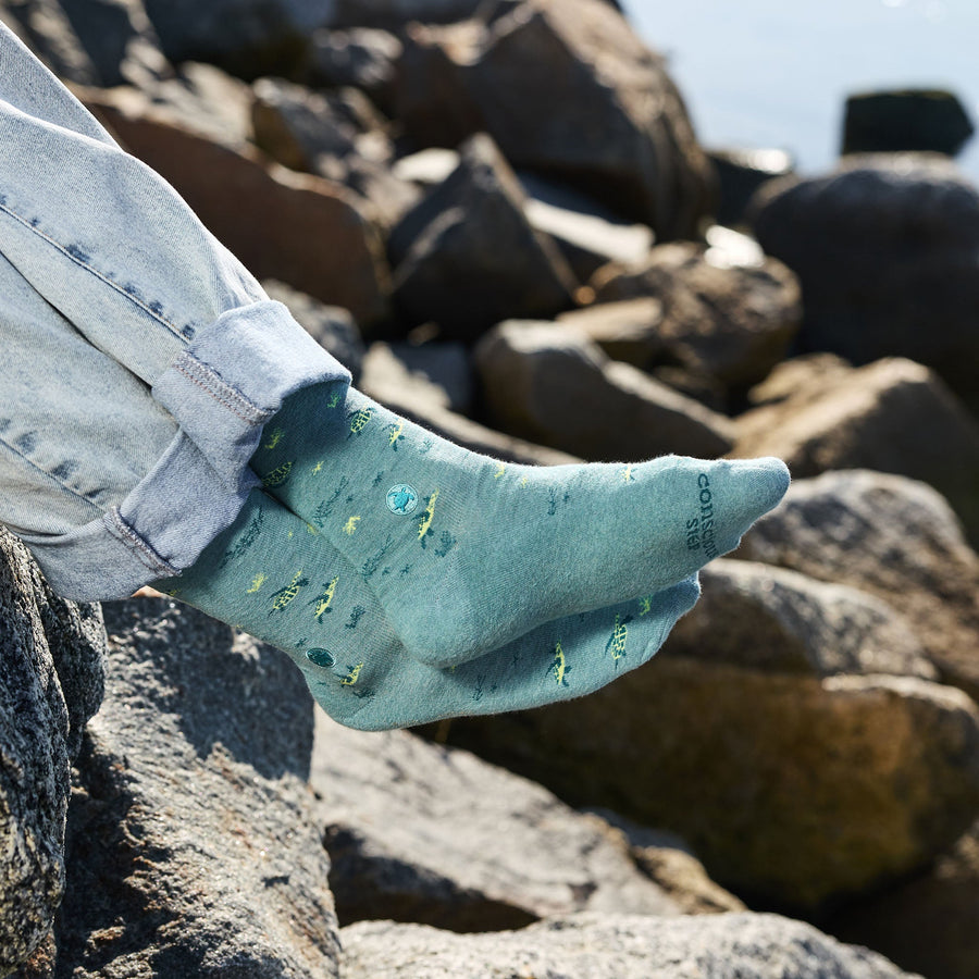 A side-profile close-up of a person's ankles and feet resting comfortably on coastal rocks, showing off the detailed turtle pattern on their teal Conscious Step socks paired with cuffed blue jeans.