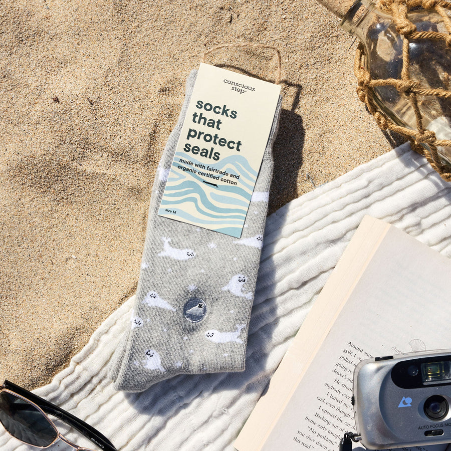 A flat lay on sandy beach featuring packaged Conscious Step gray seal socks, a white cotton towel, retro camera, open book, sunglasses, and a decorative net-wrapped glass bottle.