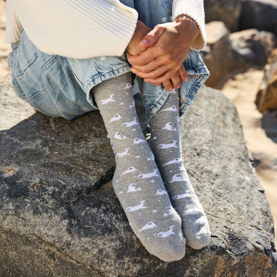 Close-up of a person's feet resting on a dark coastal rock, wearing gray crew socks with a white baby seal pattern and light-wash denim jeans.
