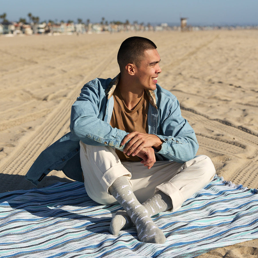 A smiling man sitting on a striped beach blanket wearing cream trousers, a brown t-shirt, a denim button-up shirt, and gray Conscious Step organic cotton seal socks.