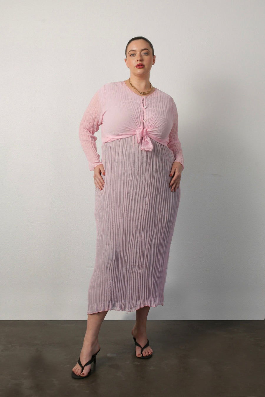 Studio portrait of a model wearing the Maya pink cardigan tied at the waist over a pleated midi dress.