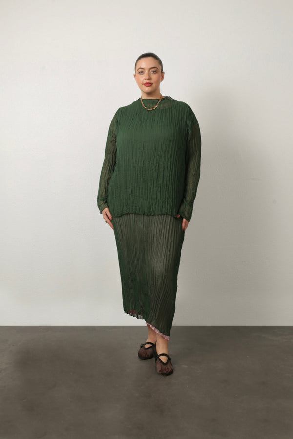 Full-length portrait of a model wearing the green Esme Pleated Funnel Neck Top styled with a matching green pleated midi skirt and gold chain necklace.