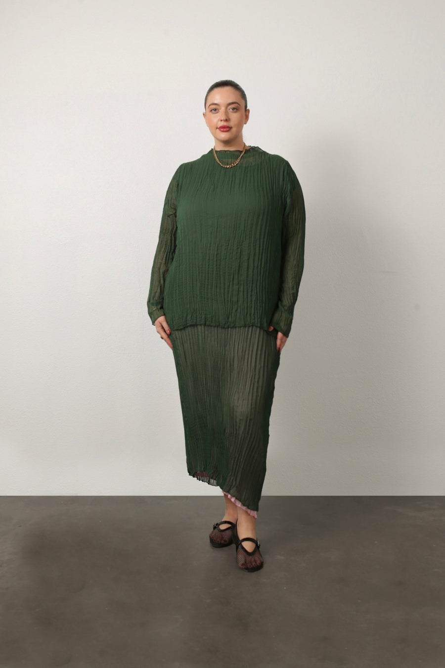 Full-length portrait of a model wearing the green Esme Pleated Funnel Neck Top styled with a matching green pleated midi skirt and gold chain necklace.
