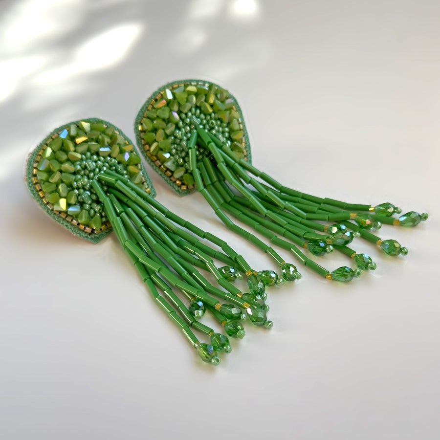 A pair of green TORTULA Moss beaded fringe earrings displayed at an angle on a white reflective surface, capturing the play of light on the iridescent faceted beads.