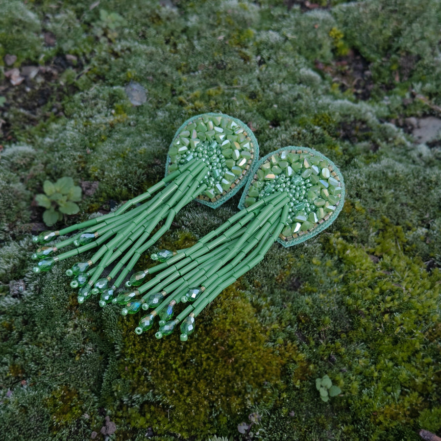 A pair of green TORTULA Moss hand-embroidered earrings laid flat on a natural bed of green forest moss, highlighting the organic texture and nature-inspired design.