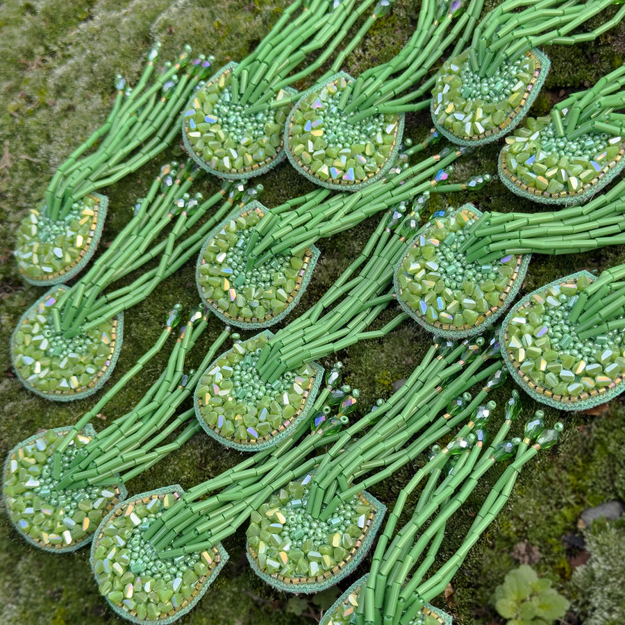 Multiple pairs of the green TORTULA Moss hand-embroidered earrings arranged together over a natural mossy surface, showcasing the repeating beaded patterns.