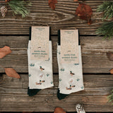 Two packaged pairs of Conscious Step Socks That Protect Ducks resting on rustic wooden planks surrounded by pine cones and autumn leaves.