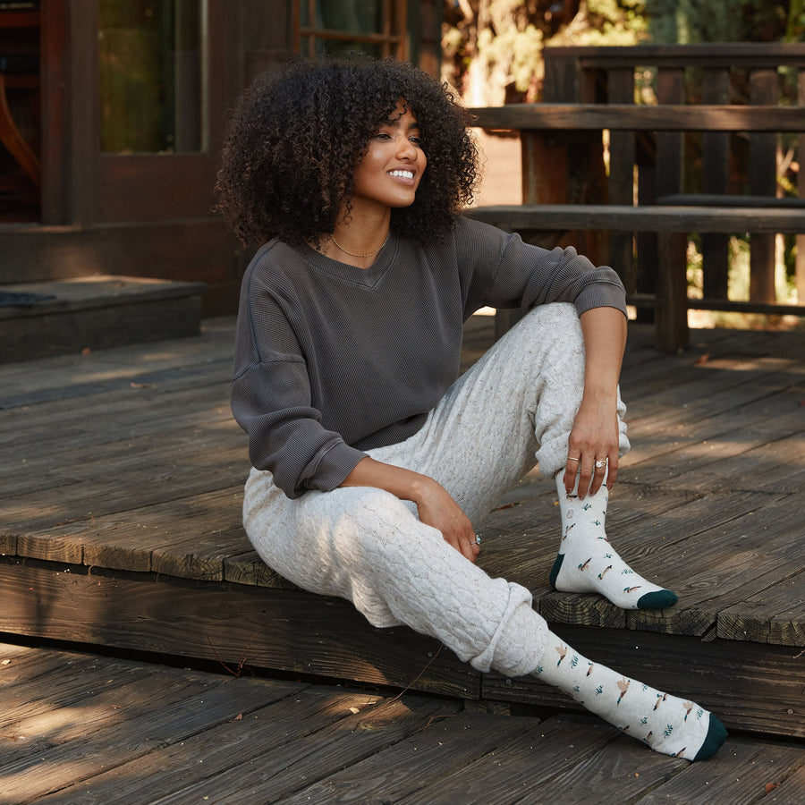 Lifestyle portrait of a woman sitting on a cabin porch wearing Conscious Step mallard duck socks paired with lounge pants and a dark sweater.