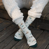 Close-up shot of feet crossed on a wooden deck showing the flying mallard duck pattern on beige organic cotton socks.