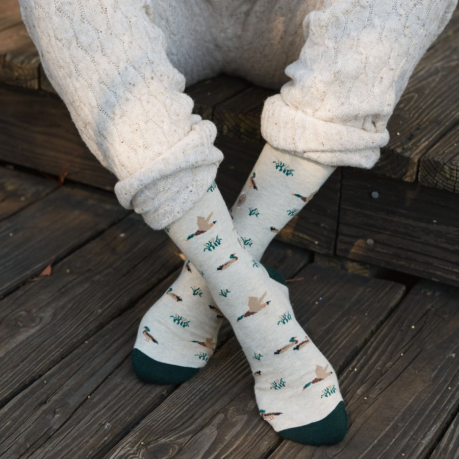 Close-up shot of feet crossed on a wooden deck showing the flying mallard duck pattern on beige organic cotton socks.