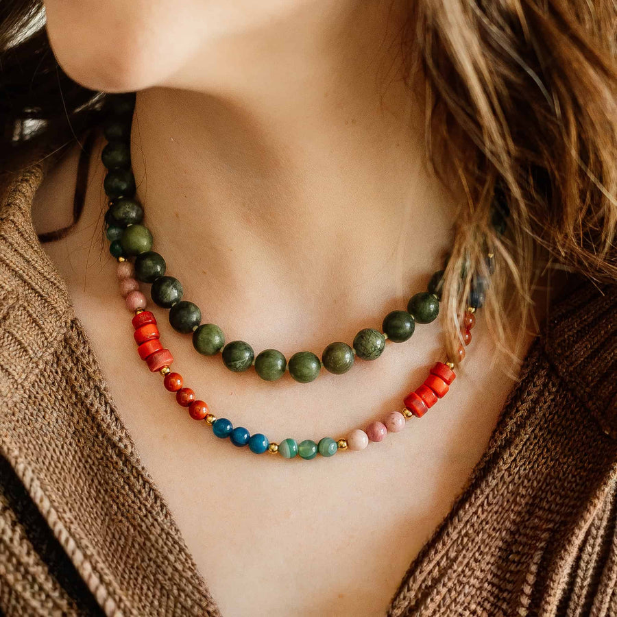 Lifestyle view of the Phoebe Gemstone Necklace layered with a dark green beaded necklace on a model wearing a brown knit sweater.