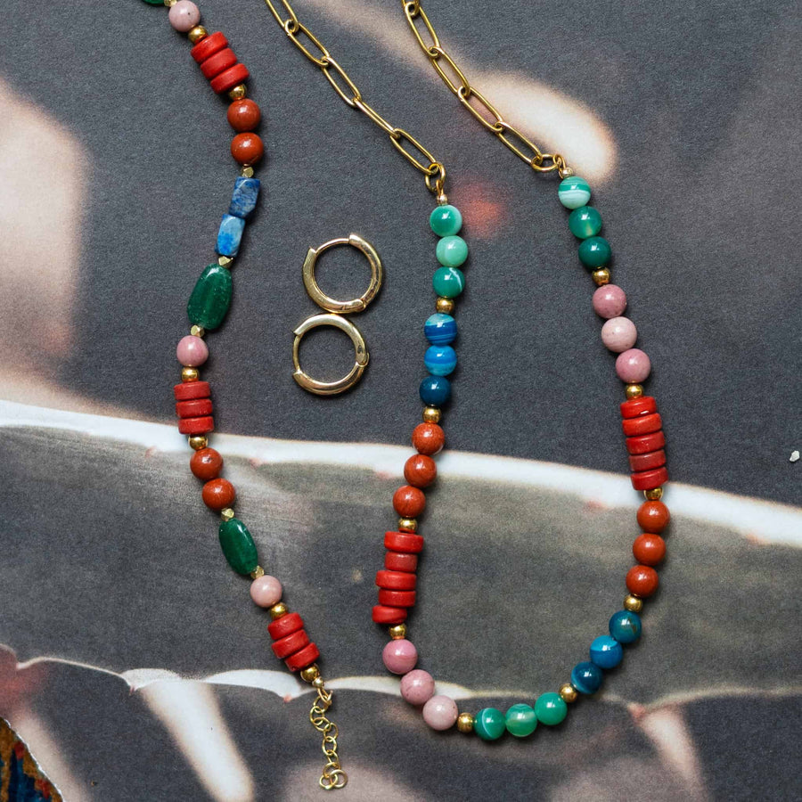 Flat lay photo of the Phoebe Gemstone Necklace alongside small gold hoop earrings on a textured background.