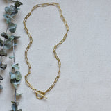 Full view of the gold Sela Designs Elle Necklace featuring a toggle closure displayed next to eucalyptus leaves.