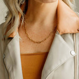 Model wearing the gold Sela Designs Elle Necklace layered over a rust-colored ribbed top and beige jacket.