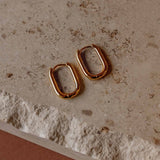 Pair of 14k gold PVD plated Sela Designs Rectangle Everyday Hoop Earrings lying on a textured neutral stone surface.