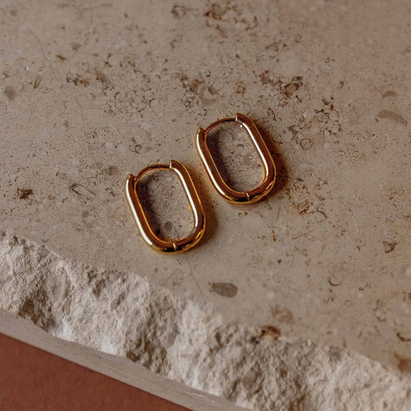 Pair of 14k gold PVD plated Sela Designs Rectangle Everyday Hoop Earrings lying on a textured neutral stone surface.