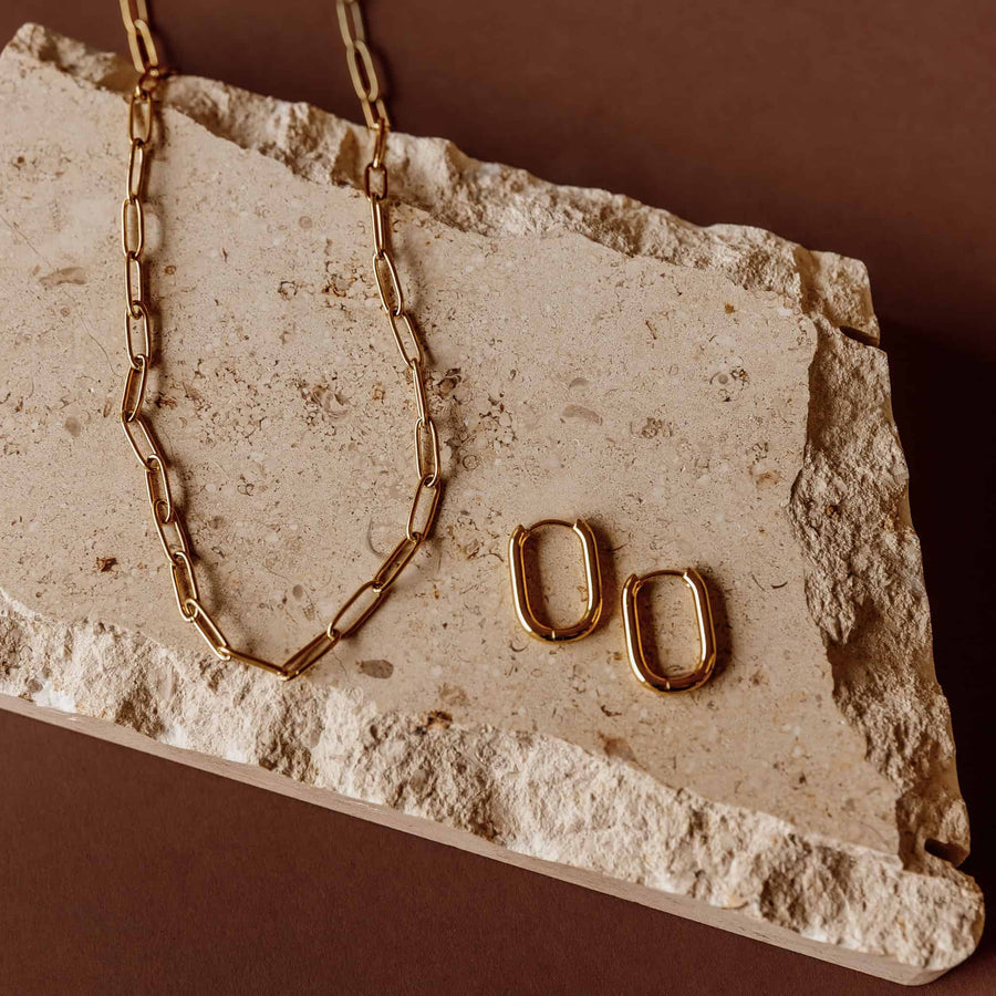 Gold Sela Designs Rectangle Everyday Hoop Earrings paired with a matching gold paperclip link chain on a stone slab.