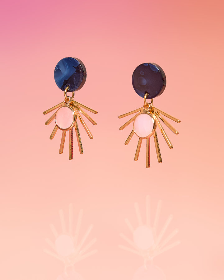 Tribe and Temple Sia Celeste Color Pop Small Dangle earrings featuring a dark navy blue marbled resin circular post, a dangling gold-plated sunburst fan, and a soft pink circular glass drop element in the center, set against a pink gradient studio background.