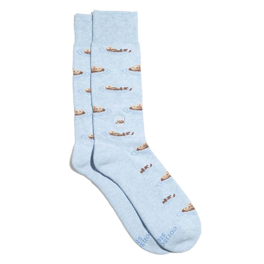 A pair of light blue crew socks from Conscious Step featuring a repeating pattern of small brown sea otters floating on water, with a round embroidered otter patch on the ankle, shot on a solid white background.