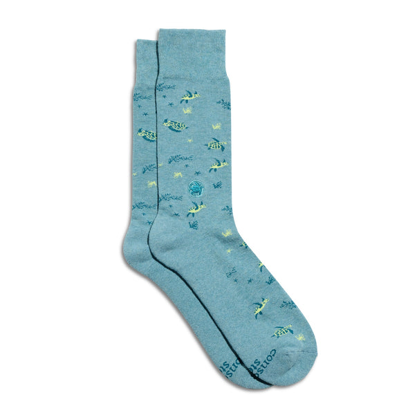 A pair of light teal crew socks from Conscious Step featuring a yellow and dark green sea turtle pattern with small underwater plants, including a circular embroidered sea turtle ankle patch, shot on a plain white background.