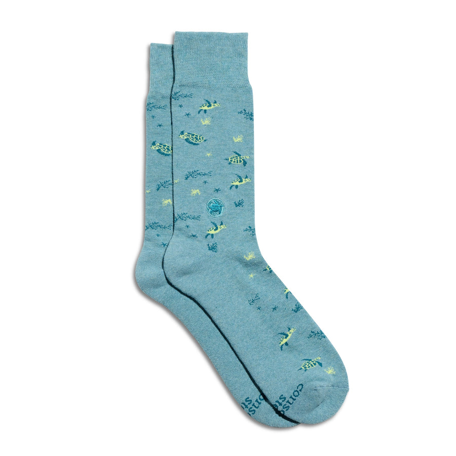 A pair of light teal crew socks from Conscious Step featuring a yellow and dark green sea turtle pattern with small underwater plants, including a circular embroidered sea turtle ankle patch, shot on a plain white background.