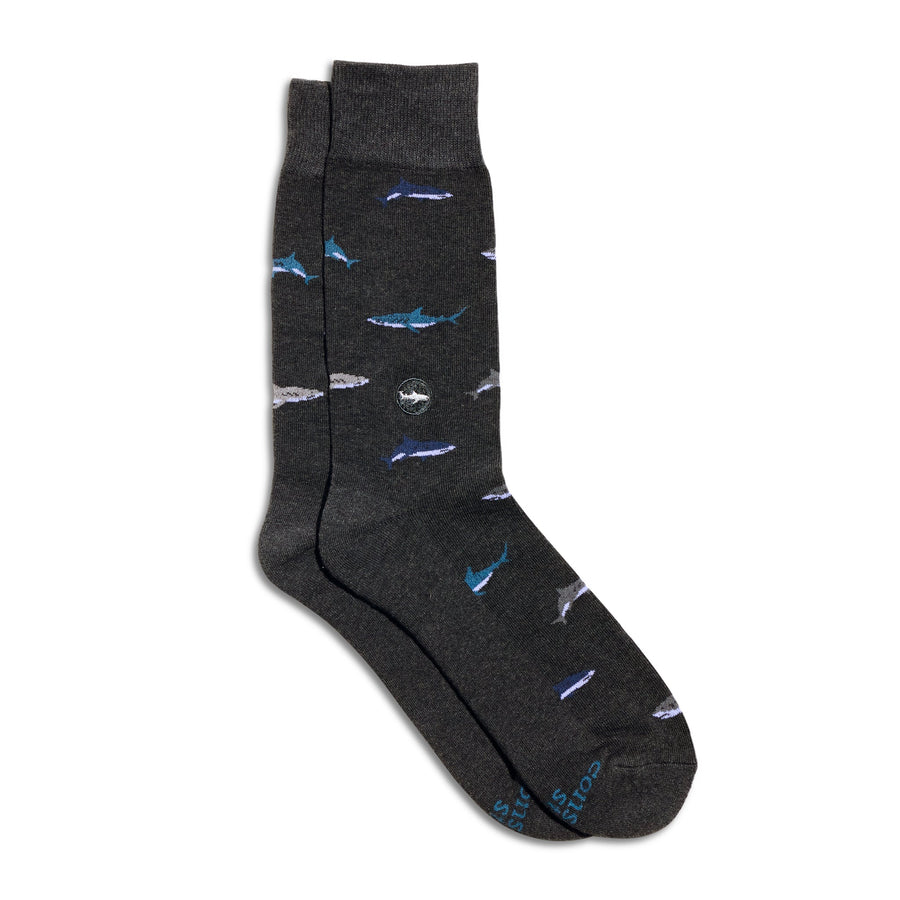 A pair of dark heather gray crew socks from Conscious Step with a repeating pattern of swimming blue and white sharks, featuring a circular embroidered shark ankle patch, shot on a plain white background.