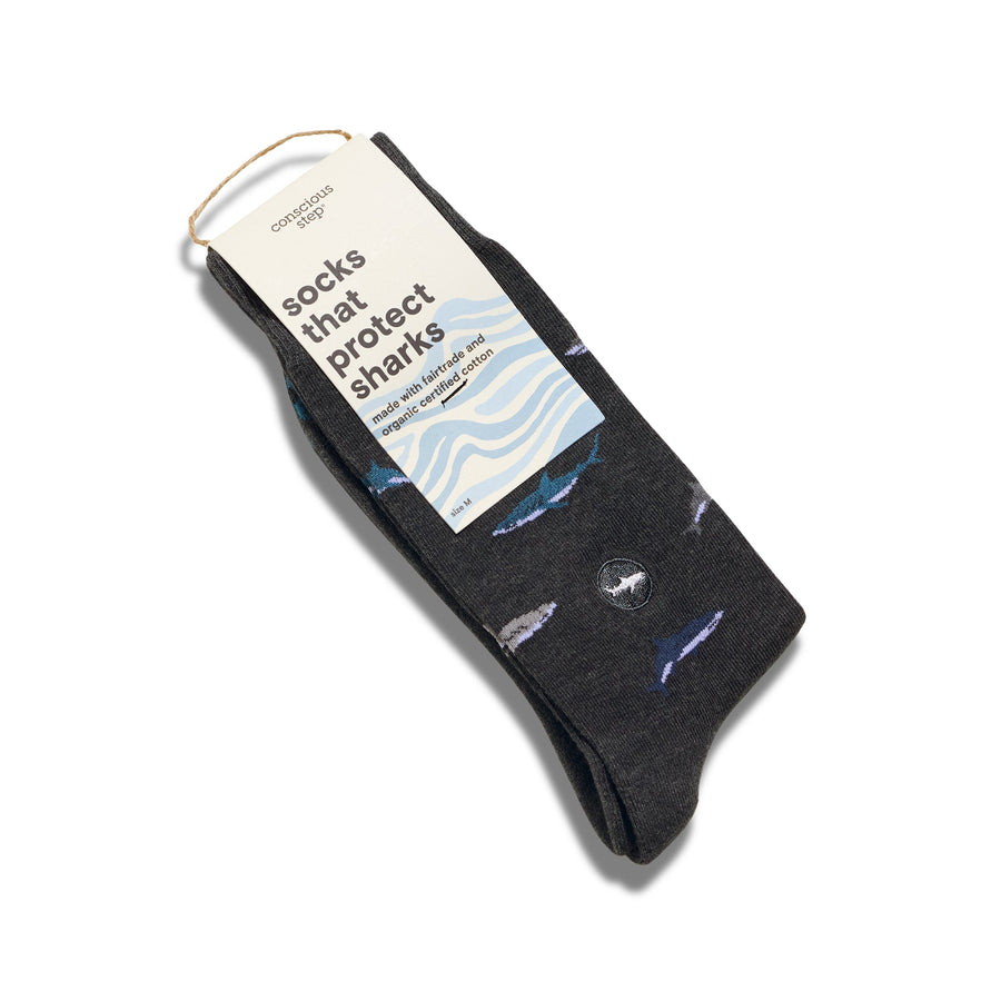 A single pair of dark gray Conscious Step shark socks folded flat with a cream card stock packaging tag that reads "socks that protect sharks made with fairtrade and organic certified cotton".